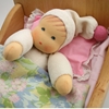 A little doll with white hat and pink pompom is lying in a bed with a pink pillow and a flowery duvet.