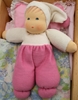Una bambolina in spugna bianca e rosa è sdraiata su un letto con un cuscino rosa e un piumone a fiori