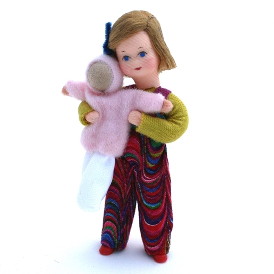 Klein meisje, poppenhuis popje met blond haar, een strikje, een veelkleurige lange broek en een groene trui. Ze draagt een roos popje in haar armen.