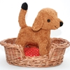 Dans un panier en osier pour chien se trouve debout un petit chien en peluche brun, debout sur le coussin à carreaux rouges et blancs qui garnit le panier.