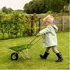 Un petit enfant en tenue de pluie se promène sur la pelouse en poussant devant lui une brouette en métal vert.